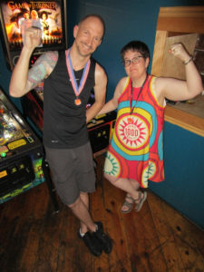 Josh and Heather both holding their fists in the air and wearing medals.  Heather has on a pinball-themed dress.