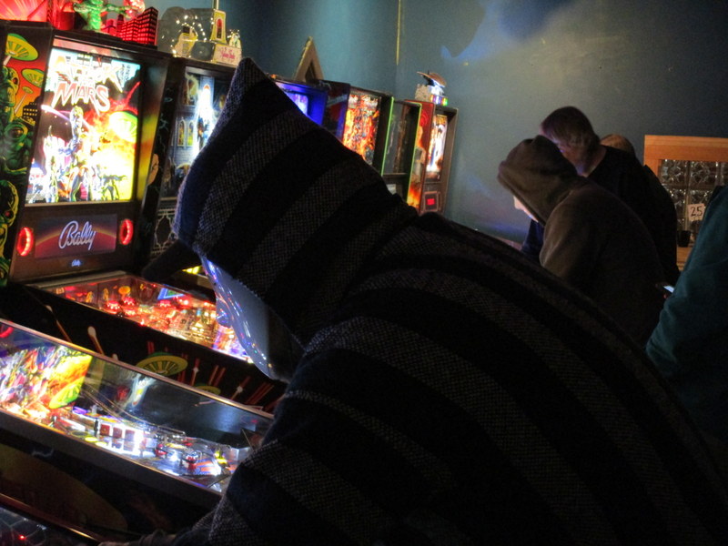 Two hooded people in hockey masks playing pinball.